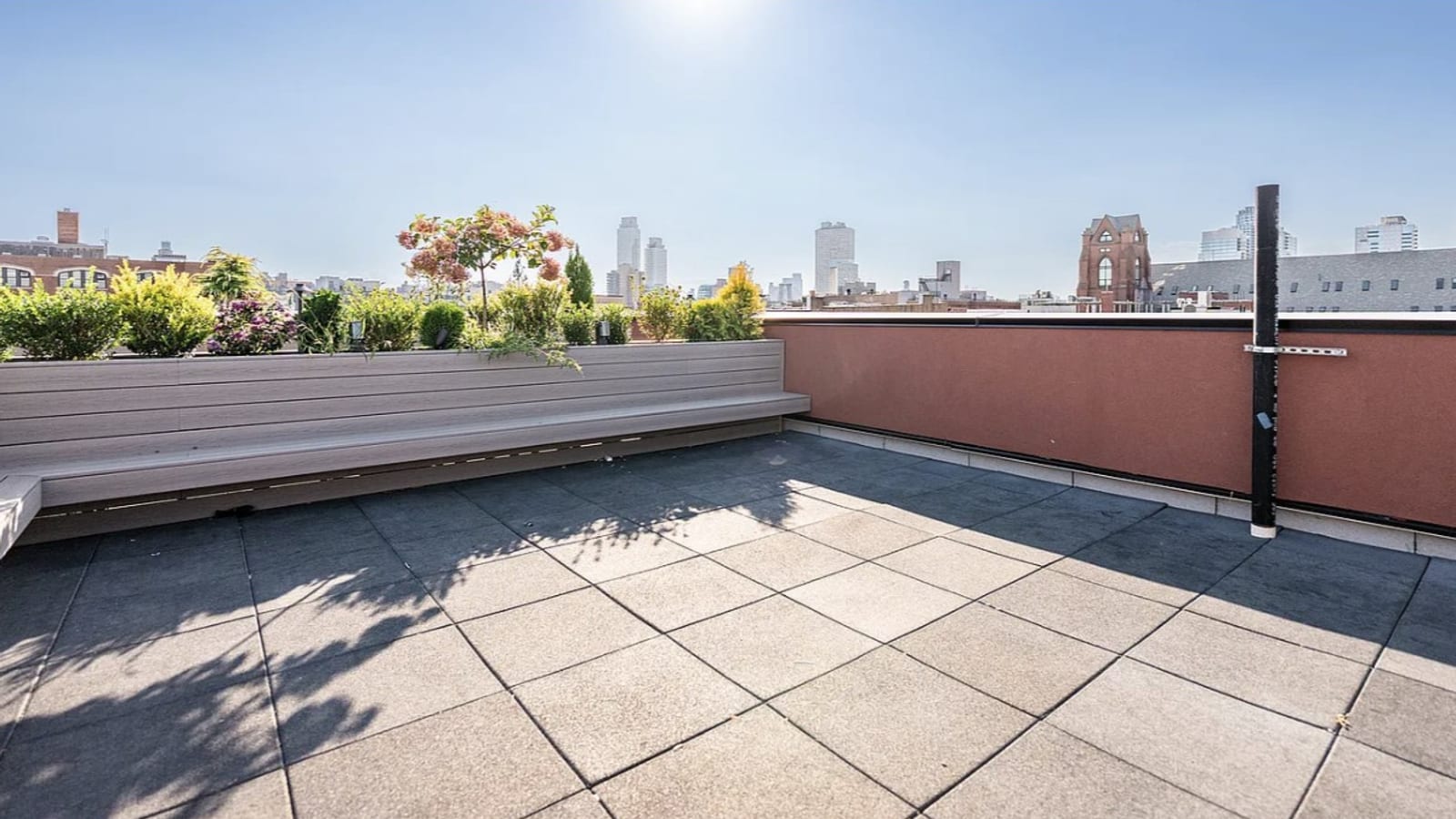 A roof terrace paved in grey tiles, with a long built-in bench and planter boxes of shrubs and a small flowering tree at left, a red parapet wall at right, and distant towers and a brick church tower on the skyline under a bright sky