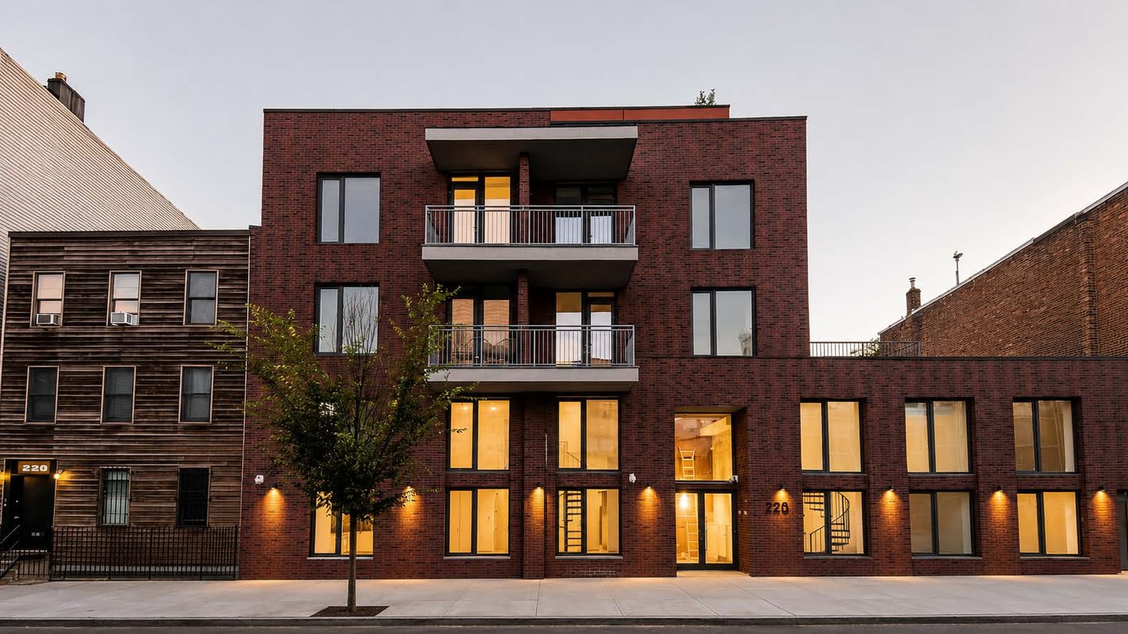 Dusk photograph of a dark red brick apartment building: a four-story section at left with two stacked balconies in the middle, a lower wing at right with a railing along its roof, tall lit ground-floor windows with spiral stairs visible inside, a glass entrance with a house number beside it, a young street tree on the sidewalk, and a wood-sided three-story house at left