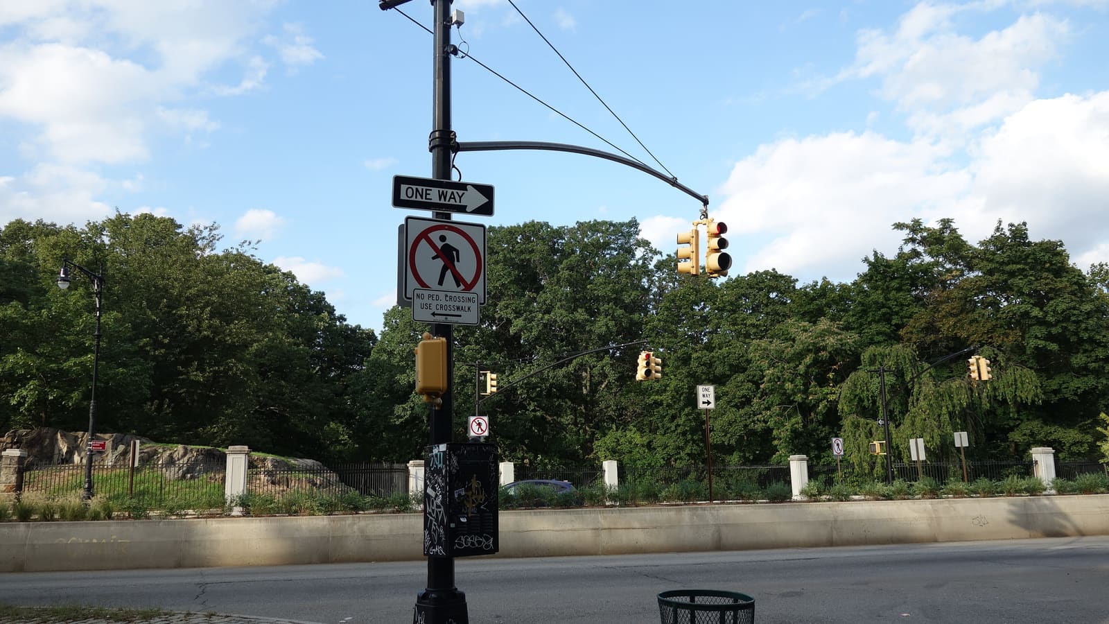 A traffic light pole with One Way and No Pedestrian Crossing signs on Fordham Road, with a stone wall, a black iron fence with white posts and tall green trees of Bronx Park beyond