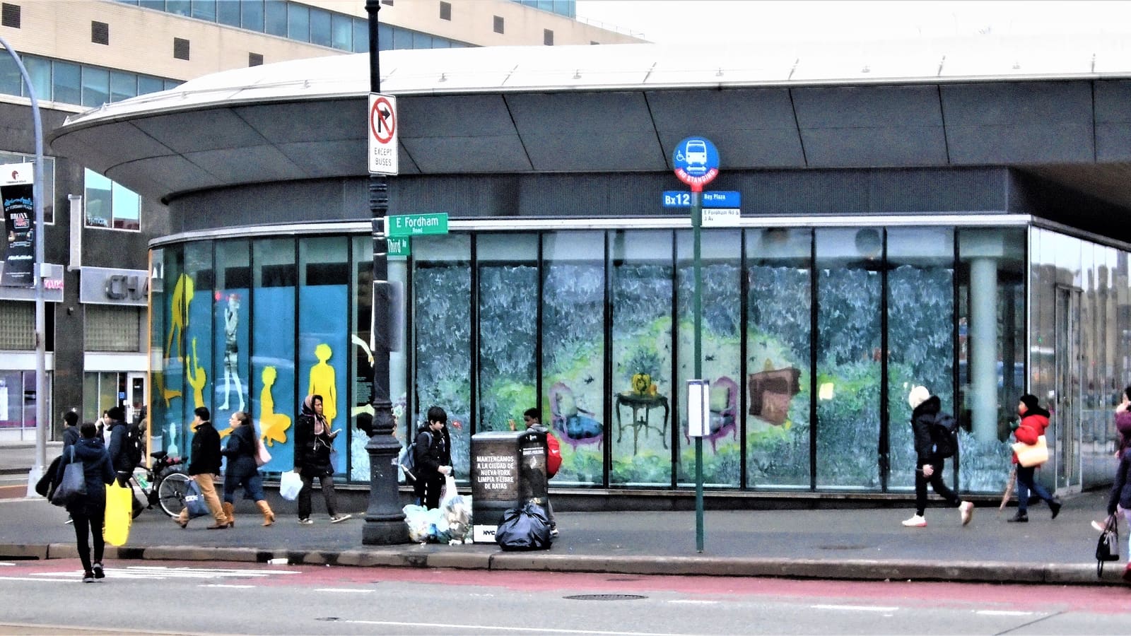 A curved glass and metal station entrance at Fordham Plaza with colorful murals behind the glass, a Bx12 bus stop sign and an East Fordham Road street sign, with people walking along the sidewalk at a distance
