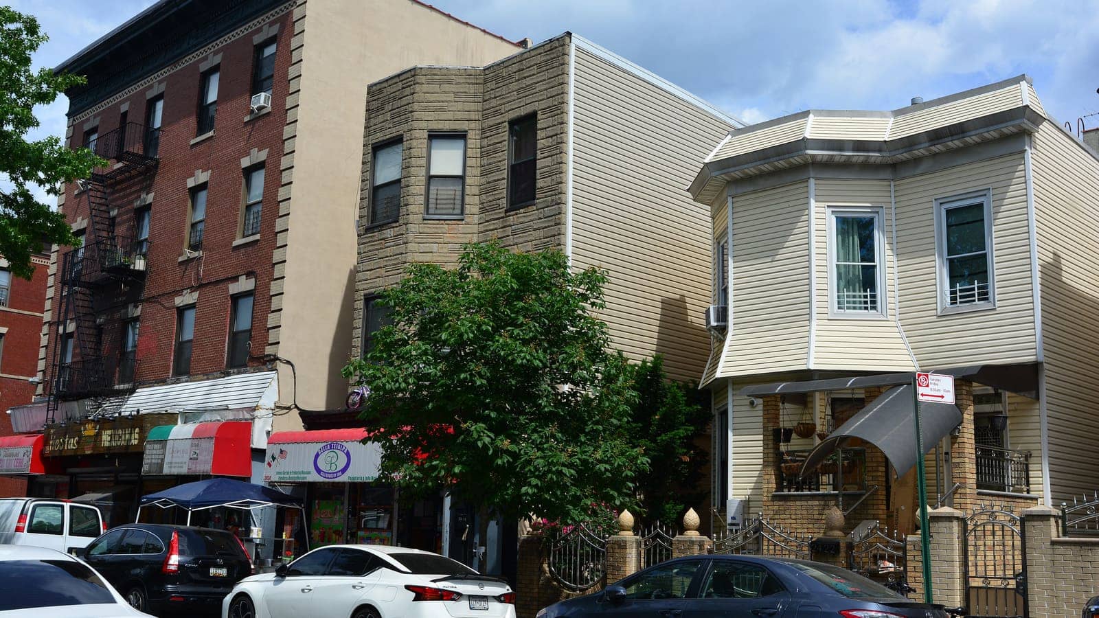 A row of buildings on Hughes Avenue: a red brick walk-up with fire escapes, a tan brick three-story building and a two-story house with cream siding and a bay window, with shop awnings, a street tree, a gate and parked cars in front