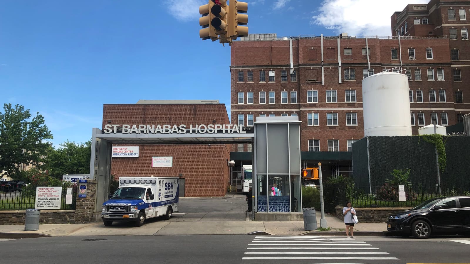 The St Barnabas Hospital emergency entrance with its name in large letters over a driveway, an ambulance at the curb, a red brick hospital building and a white tank behind, traffic lights and a crosswalk in front