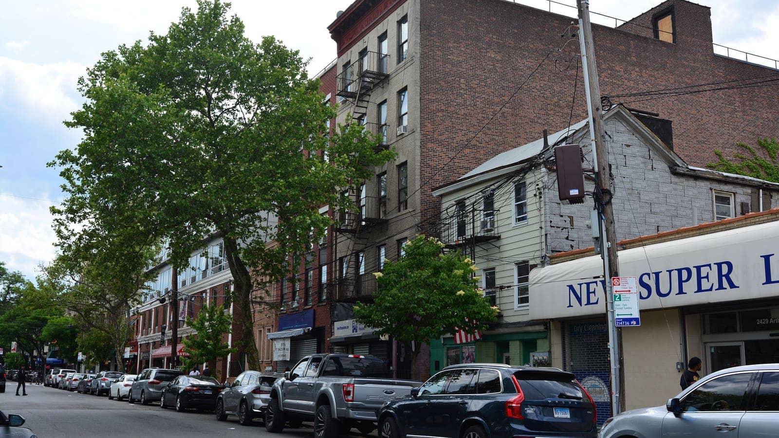A tree-lined block of Arthur Avenue with four- and five-story brick apartment buildings with fire escapes, a white-sided older house, a grocery awning reading New Super, and a row of parked cars under a cloudy sky