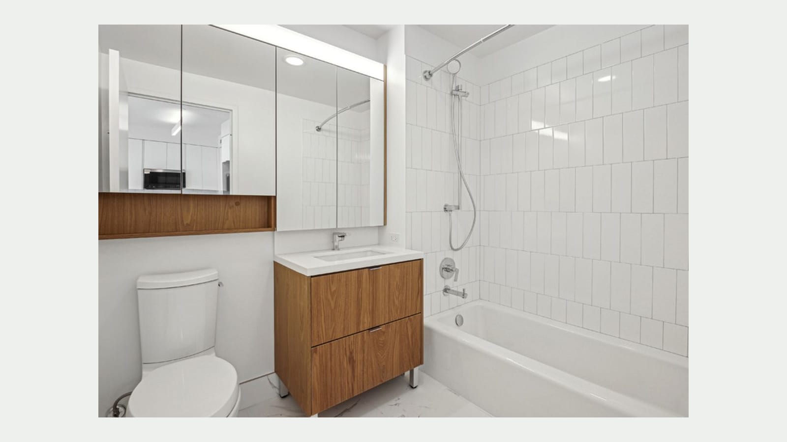 Photograph of a bathroom with a wood vanity and white sink, a wide mirrored cabinet over a wood shelf, a white toilet, a tub with white vertical tile and a hand shower, and a white marble-look floor