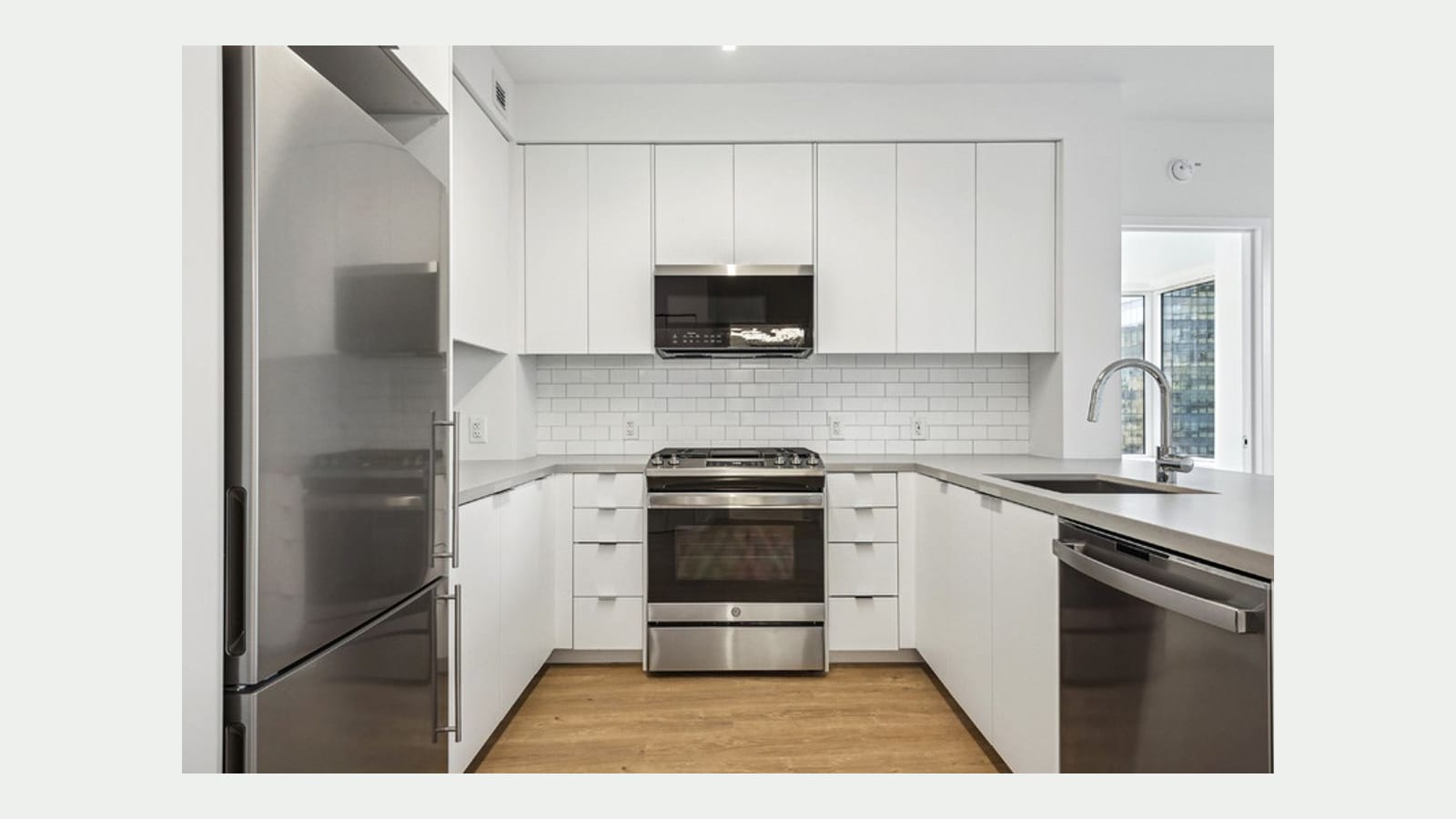Photograph of an empty U-shaped kitchen with flat white cabinets, a white subway tile backsplash, a stainless gas range with a microwave above, a stainless refrigerator at left, a dishwasher and sink at right, gray counters, light wood floors and a window to other towers