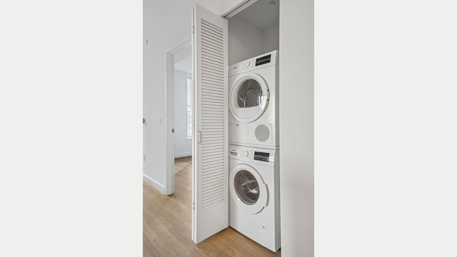 Photograph of a stacked white Bosch washer and dryer in a closet with a louvered white folding door, beside a doorway to an empty room with a window and light wood floors