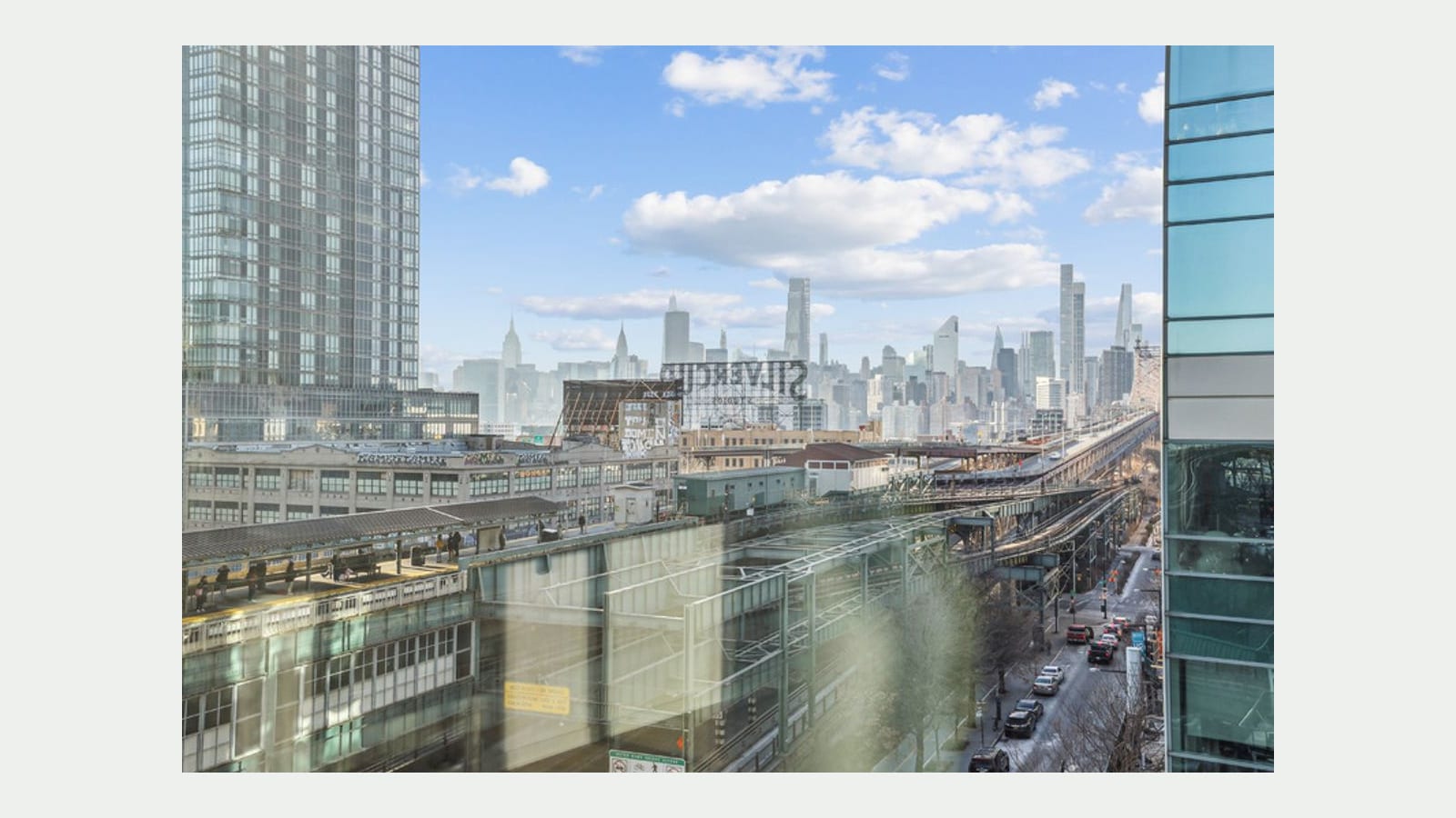 Photograph from a window high in the building looking over elevated train platforms and tracks, the back of the Silvercup sign, low industrial buildings and a street with parked cars, with the Manhattan skyline beyond, a glass tower at left and a pale glare streak on the window