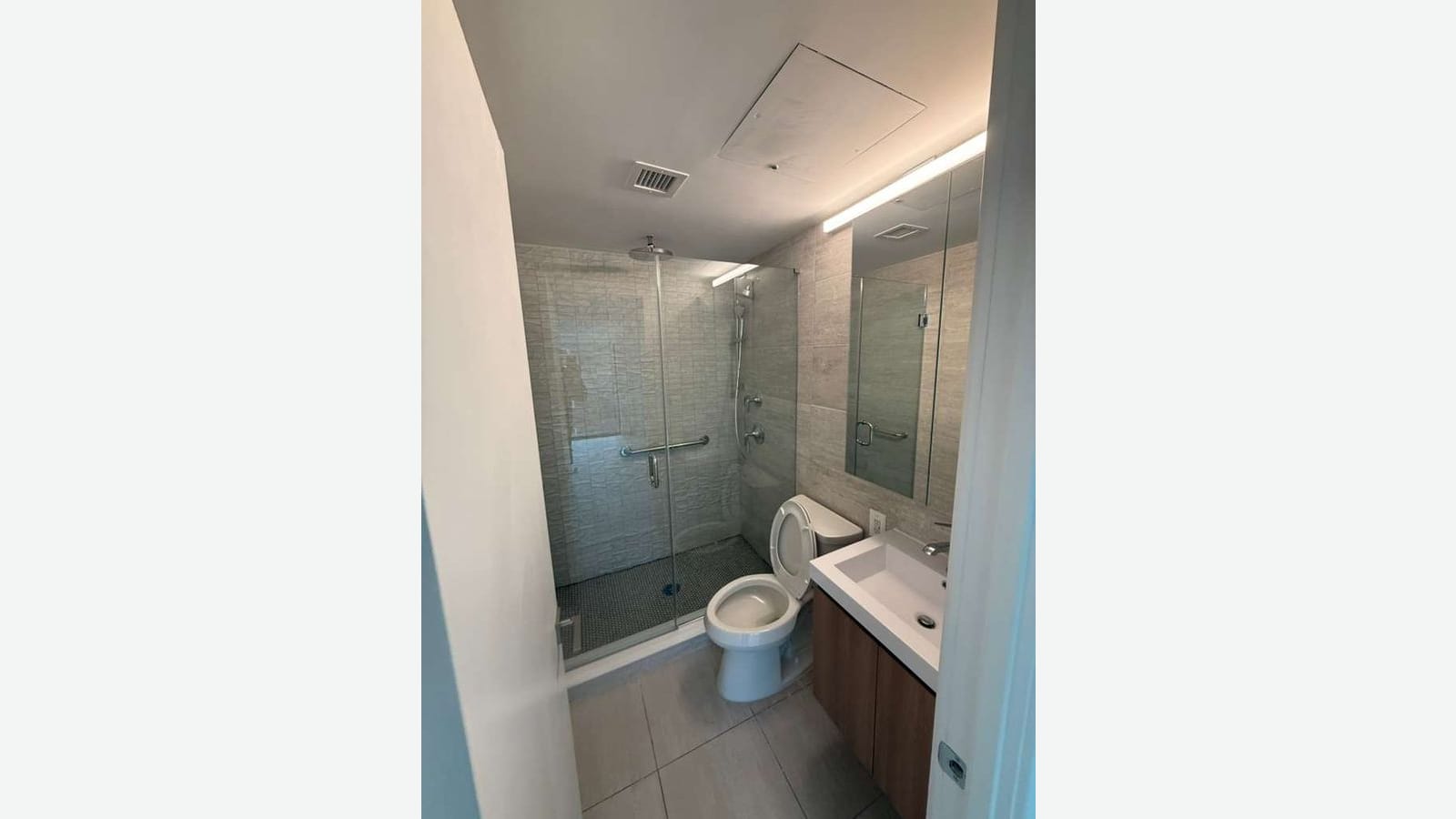 Photograph of a bathroom with a glass-walled shower, gray textured wall tile, a rain shower head and grab bar, a white toilet, a white sink on a wood-grain vanity, a mirror cabinet and a light bar