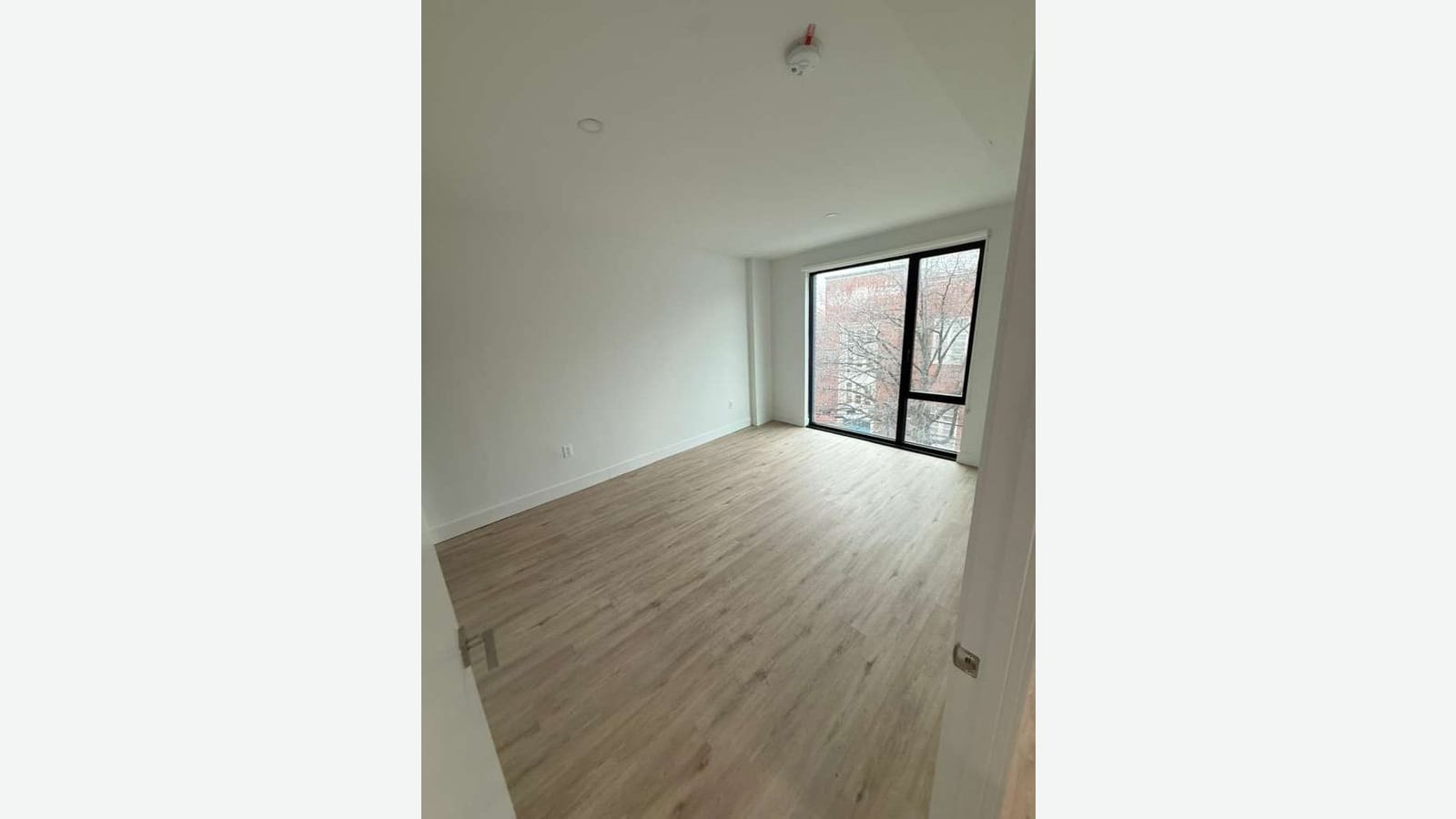 Photograph of an empty bedroom with white walls, wood-look plank floors, a smoke detector on the ceiling, and a tall black-framed window looking out on bare trees and a brick building
