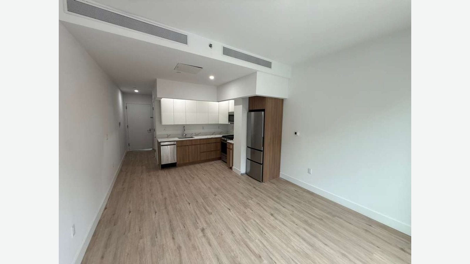 Photograph of an empty studio with wood-look plank floors, white walls, a ceiling air vent, and a kitchen at the far end with white upper cabinets, wood-grain lower cabinets, a stainless steel dishwasher, a range and a stainless steel refrigerator, next to the apartment door