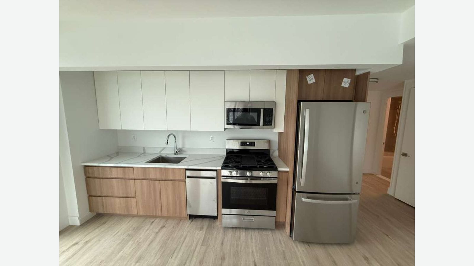 Photograph of a kitchen wall with white upper cabinets, wood-grain lower cabinets, a white stone counter with a sink, a stainless steel dishwasher, a stainless steel range with black burner grates, a microwave above it and a stainless steel refrigerator