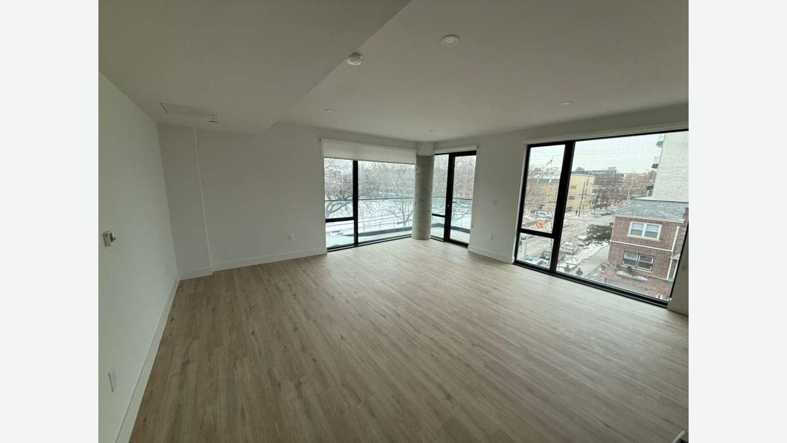 Photograph of an empty corner living room with wood-look floors, floor-to-ceiling windows on two walls, a glass door to a balcony, and a view of bare trees, a street and a brick house in winter