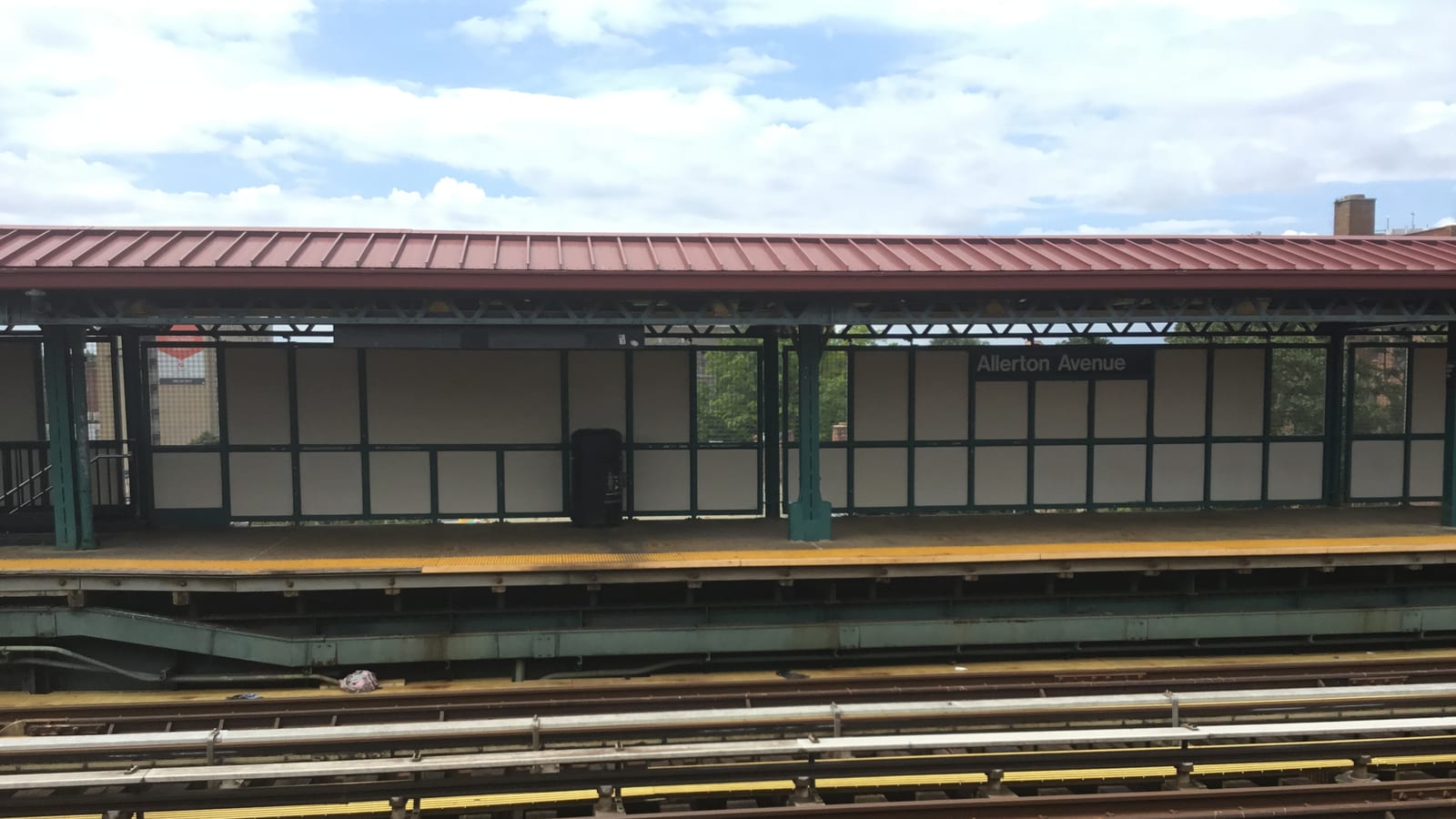 Photograph of an outdoor subway platform with a red metal canopy, green steel columns, a sign reading Allerton Avenue, a yellow platform edge and several tracks in the foreground under a partly cloudy sky