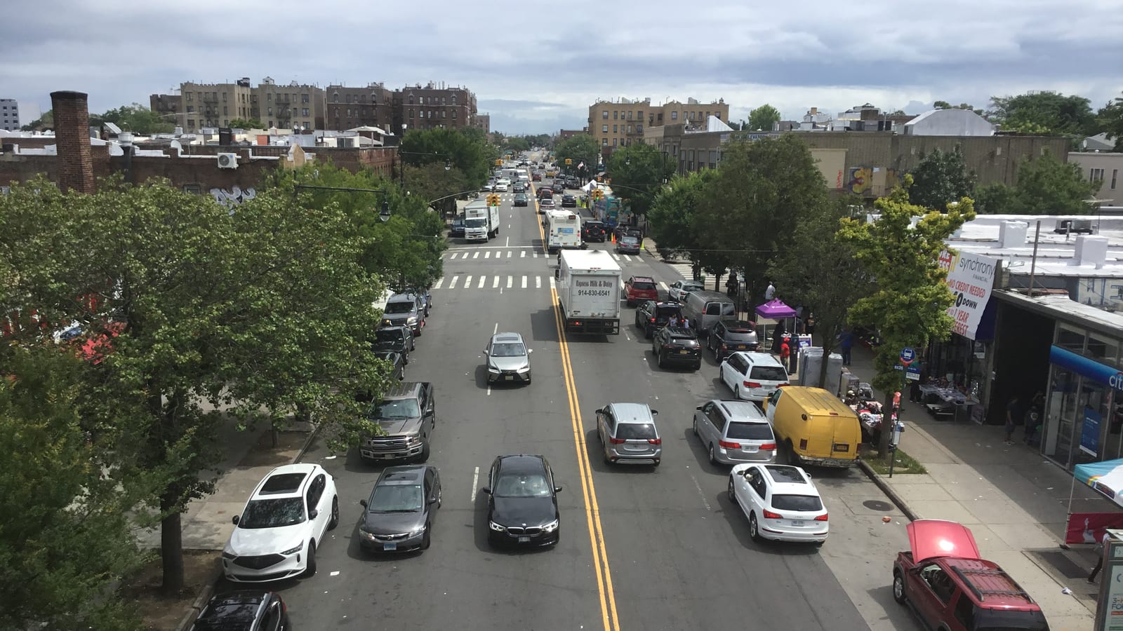 Photograph looking down a busy two-way avenue from above, with parked and moving cars, a box truck, street trees on the left, shops with awnings on the right and low brick apartment buildings in the distance