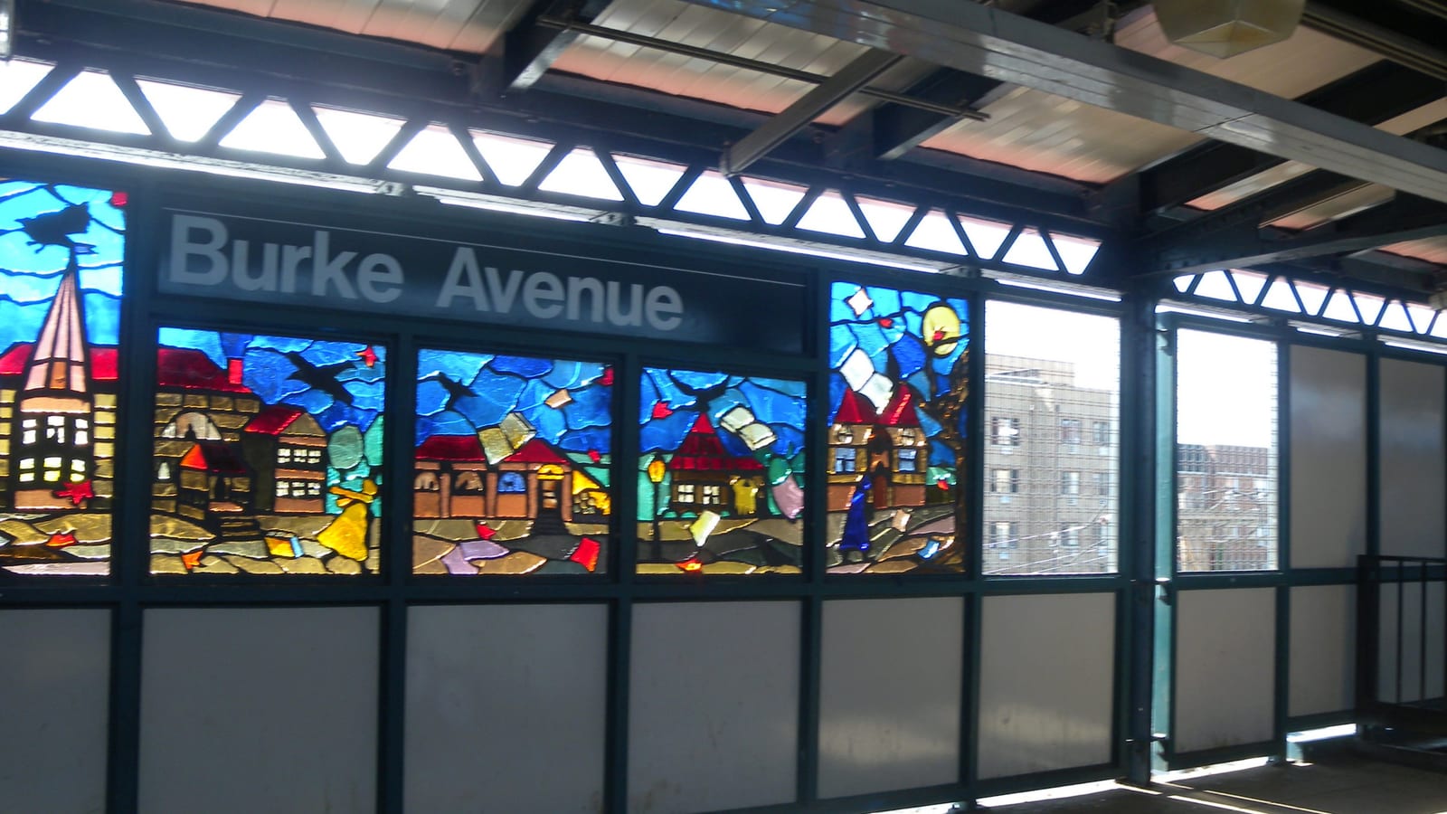 Photograph of a subway platform windscreen with colorful stained-glass panels showing houses, birds and flying books, under a sign reading Burke Avenue, with sunlight casting colored light on the platform floor