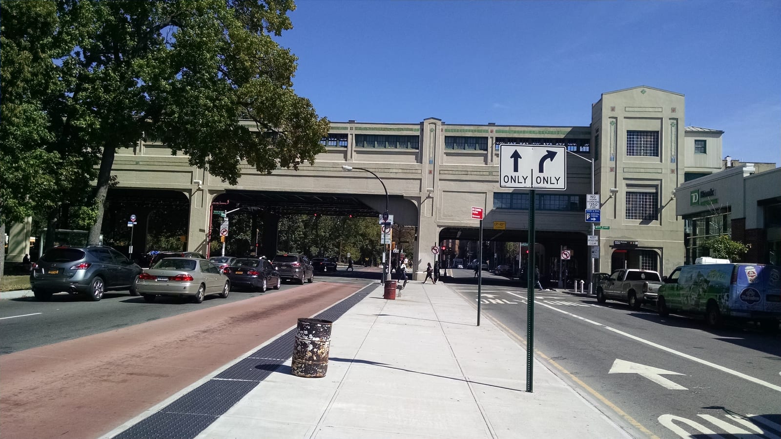 Photograph of a wide street with a red bus lane and a sidewalk leading to a long stone-colored station building that spans the road on a steel structure, with a lane-direction sign, parked cars and leafy trees under a blue sky