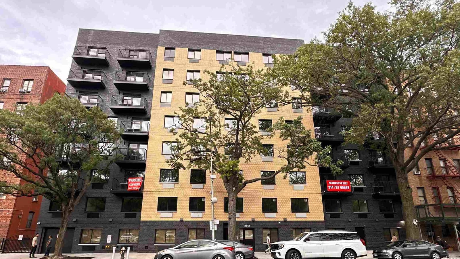 Photograph of an eight-story apartment building with a tan brick center section and dark gray brick wings, black metal balconies on the wings, two red signs reading Parking for Rent, the number 2512 over a glass entrance door, street trees in front, cars parked at the curb and older brick apartment buildings on both sides