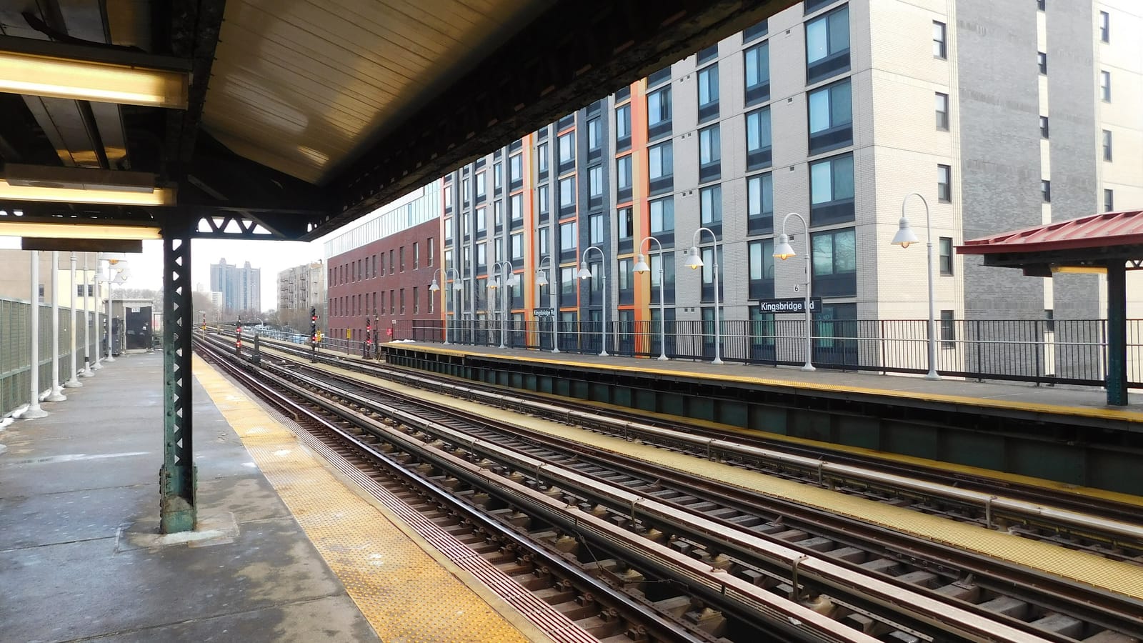 Photograph from a covered subway platform above the street, looking across four tracks to a second platform with white lamp posts and a sign reading Kingsbridge Rd, with a tall pale gray apartment building with orange trim and a lower red brick building behind it under a pale sky