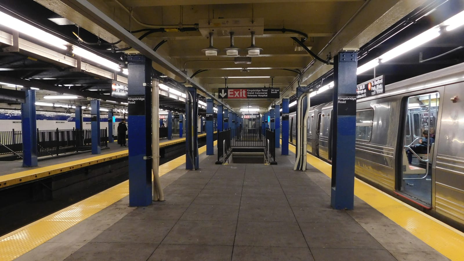 Photograph of an underground subway platform with blue columns labeled Kingsbridge Road, yellow edge strips, a stair down in the middle under a red Exit sign naming Kingsbridge Road, Grand Concourse and Veterans Hospital, and a silver train with open doors at right