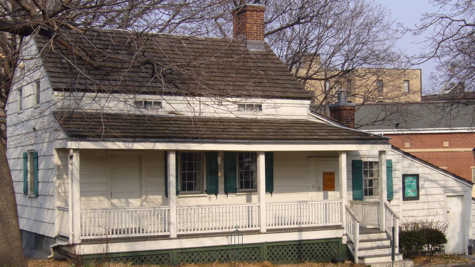 Photograph of a small white wooden farmhouse with a brown shingle roof, a red brick chimney, green shutters and a long front porch with a white railing, behind bare tree branches, with brick buildings in the background