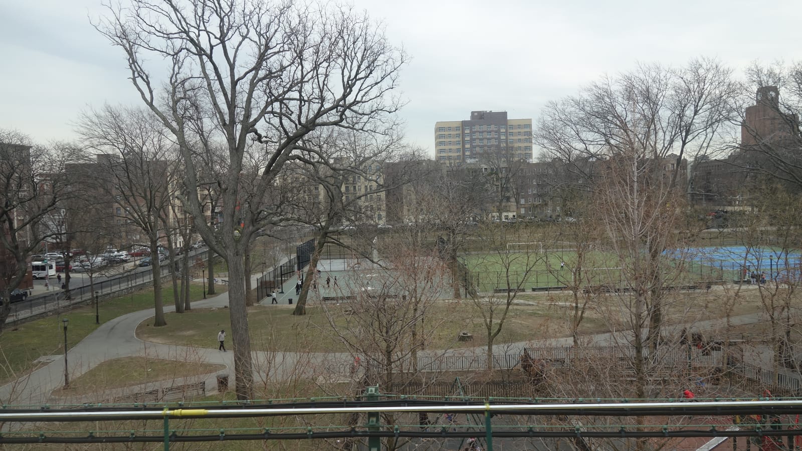 Photograph from a train above a park in early spring, with tall bare trees, curving paved paths, a basketball court, a green soccer field and blue courts, a row of parked cars along a street at left and brick and pale apartment buildings behind under a gray sky
