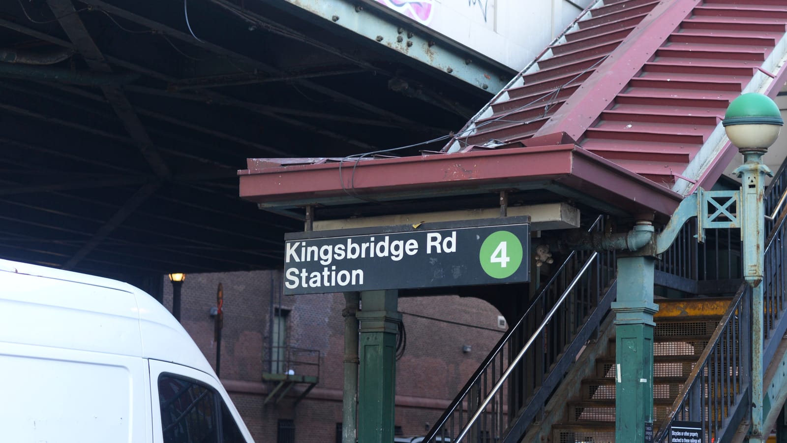 Photograph of a subway station stair under a steel structure, with a black sign reading Kingsbridge Rd Station and a green circle with the number 4, a dark red roof over the stair, green iron columns and railings, a white globe lamp at right, and the side of a white van at left in front of a brick building