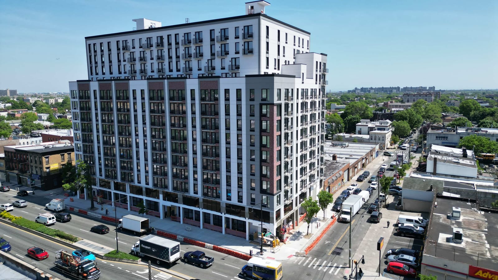 Aerial photograph of a 14-story white apartment building with dark window frames, red brick panels and small balconies on its upper floors, a setback near the top and a glass-fronted ground floor, on a corner of a wide avenue with a planted median, trucks and cars; low-rise buildings and trees stretch to the horizon under a clear blue sky
