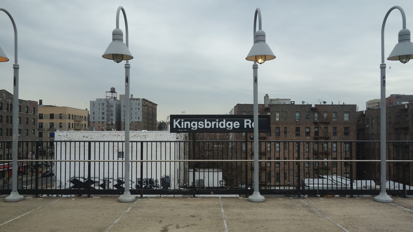 Photograph of an outdoor subway platform with four curved grey lamp posts, a black railing and a black sign reading Kingsbridge Rd, with brick apartment buildings and a white painted wall behind under a cloudy sky
