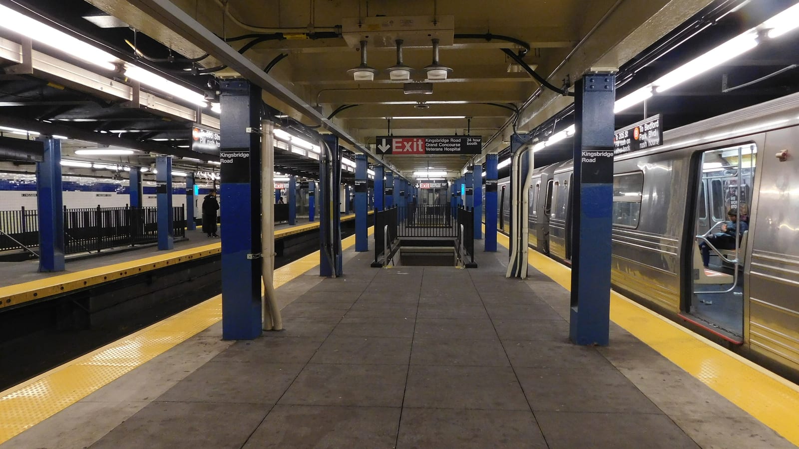 Photograph of an underground subway island platform with blue steel columns marked Kingsbridge Road, yellow platform edges, a red Exit sign naming Kingsbridge Road, Grand Concourse and Veterans Hospital, and a silver train with open doors on the right