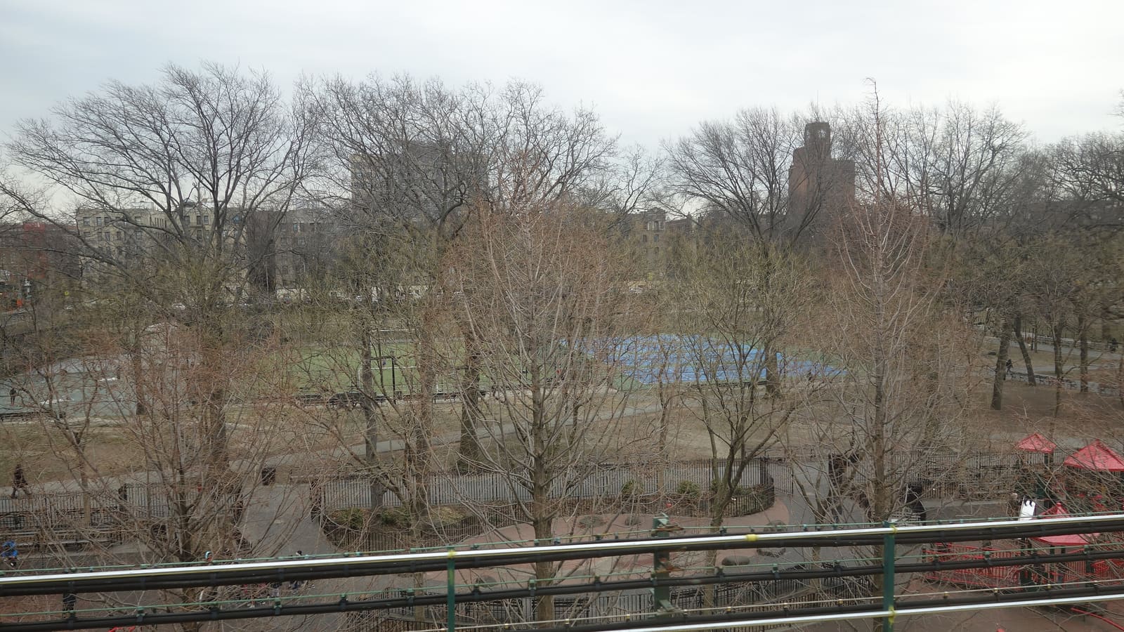 Photograph from a raised subway track of a park in early spring, with bare trees, a playground with red canopies in front, ball courts with green and blue surfaces in the middle, and apartment buildings and a brick clock tower behind