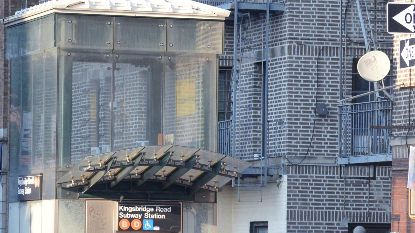 Photograph of a glass and steel subway elevator kiosk with a curved glass canopy and a sign reading Kingsbridge Road Subway Station with B and D route symbols and a wheelchair symbol, beside a brick apartment building with fire escapes