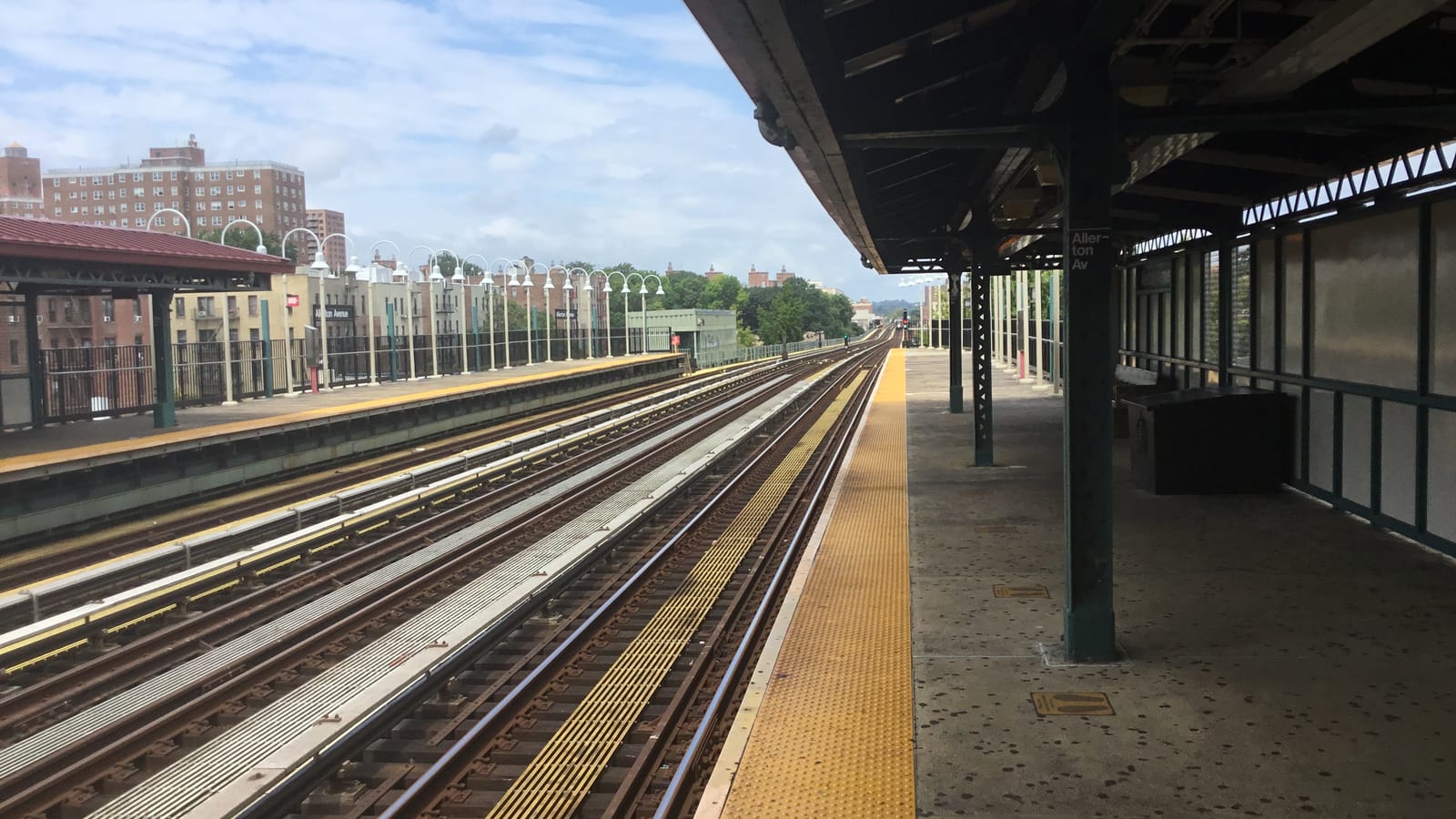 Photograph from a raised subway platform with a green steel canopy and a sign reading Allerton Av, looking along three tracks toward the far platform with white lamp posts, and brick apartment towers and trees behind under a blue sky
