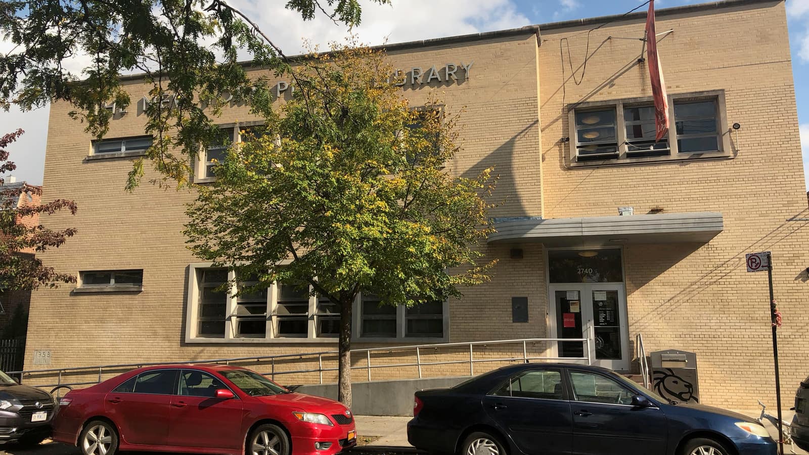 Photograph of a two-story tan brick library with a curved metal canopy over a glass door numbered 2740, a red banner on a pole, raised lettering reading The New York Public Library partly hidden by a street tree, a ramp with railings, and a red car and a blue car parked in front