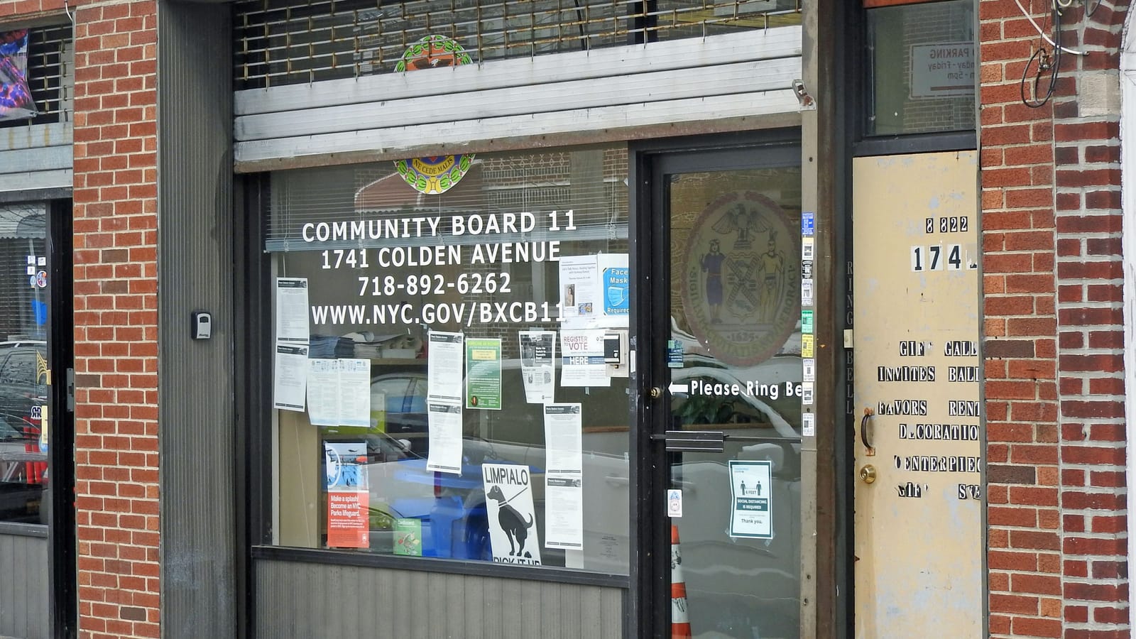Photograph of a brick storefront office with a large window lettered Community Board 11, 1741 Colden Avenue, a phone number and a web address, notices taped inside the glass, a City seal on the glass door, and a half-raised metal gate above