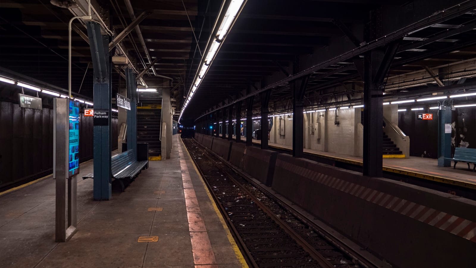 Photograph of an empty covered subway platform with a bench, a digital information screen, a sign reading Pelham Parkway on a blue column, stairs up to the street, and a single track beside a low concrete wall, with a second platform across the tracks