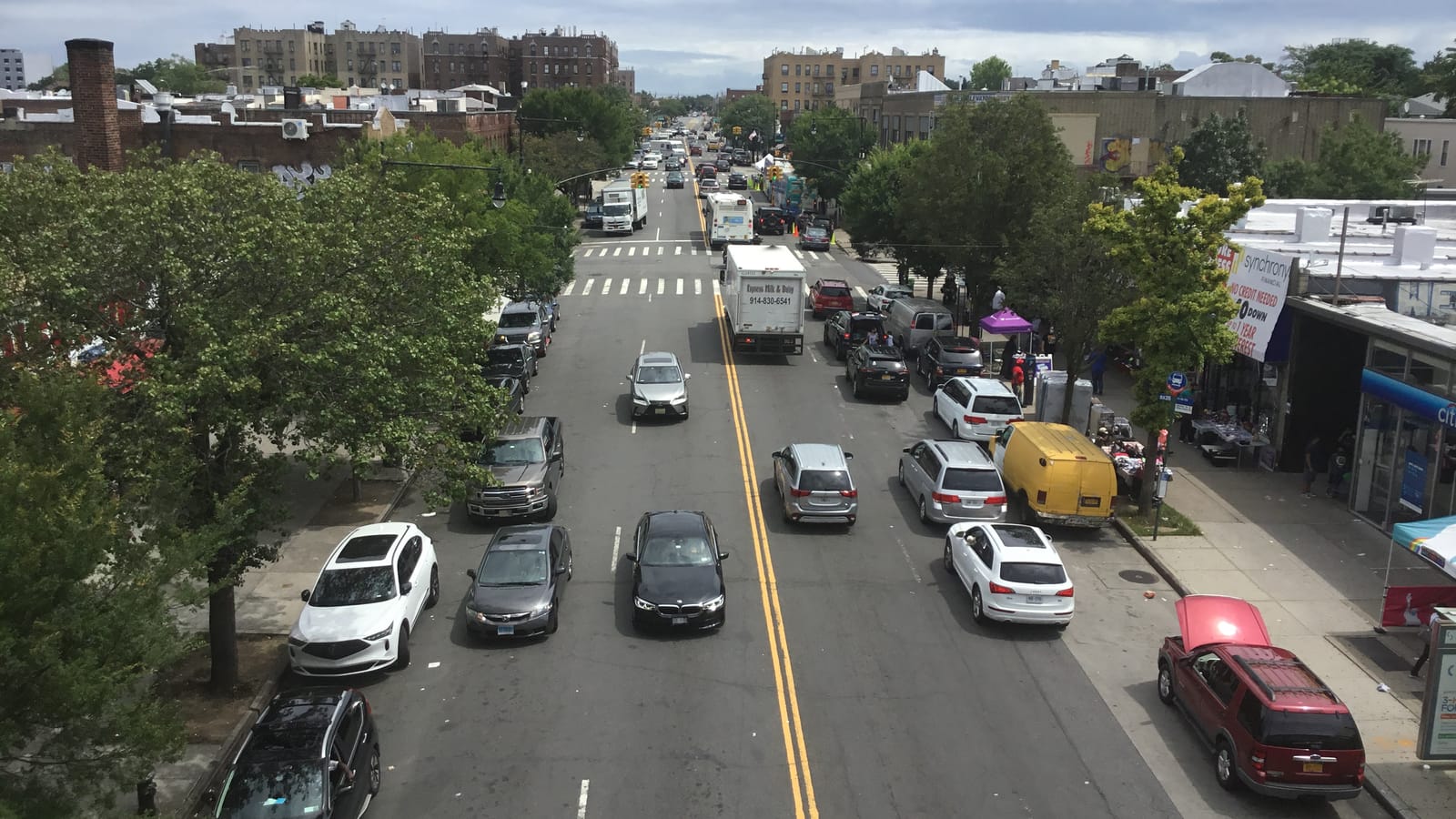 Photograph looking down a wide two-way street lined with trees and parked cars from a raised subway platform, with a box truck and a bus in traffic, shops and a bank branch on the right, and brick apartment buildings on the horizon under a cloudy sky