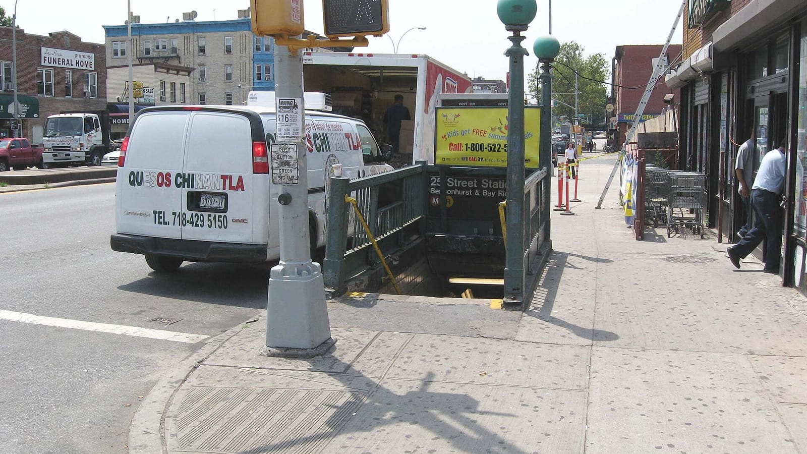 Green iron subway stair entrance with two globe lamps on a wide sidewalk, a 25 Street Station sign, a pedestrian signal pole at the corner, a white delivery van at the curb and low brick buildings with a funeral home sign across the avenue