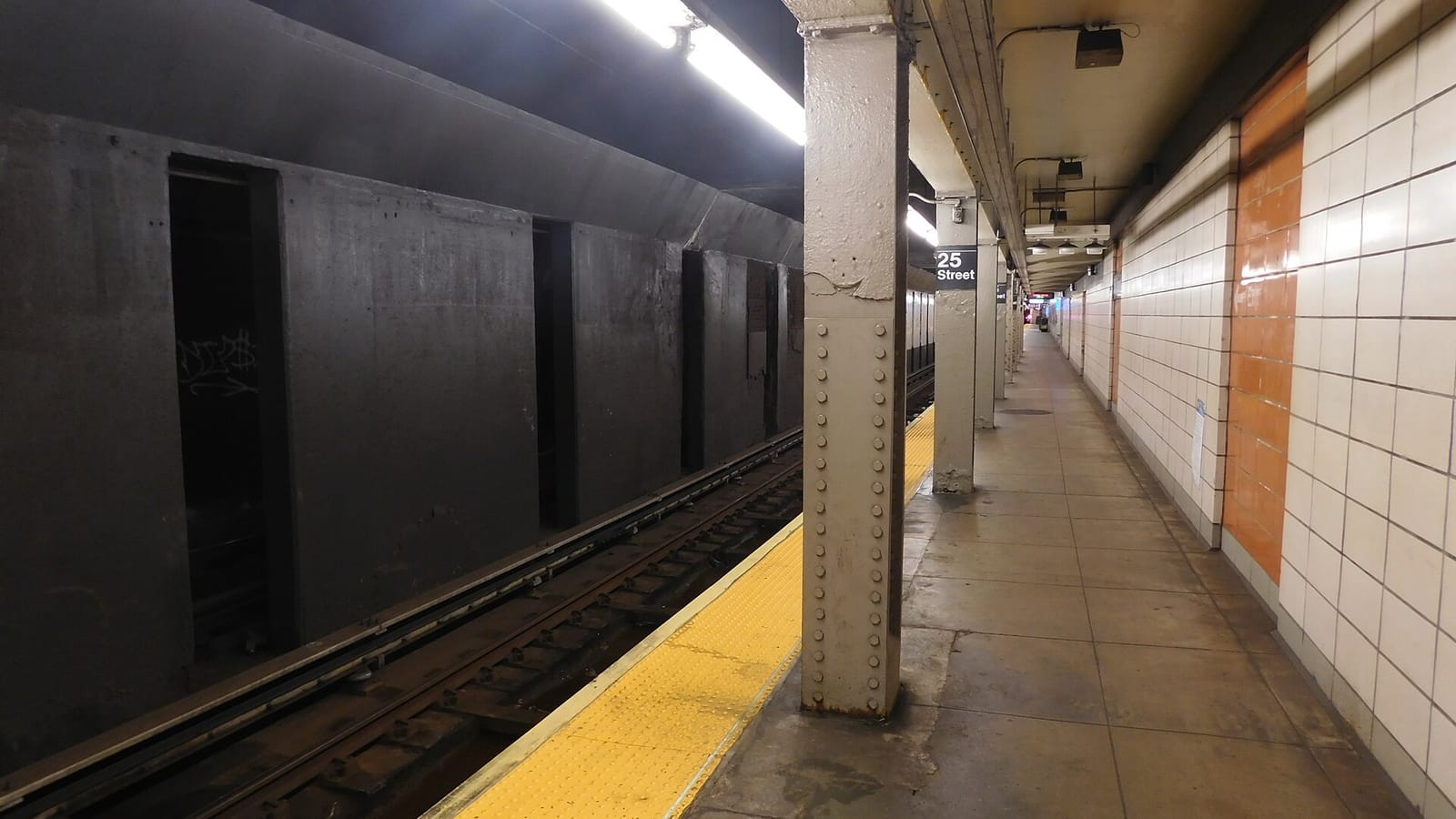 Empty underground subway platform with riveted steel columns, a yellow edge strip, white wall tiles with an orange band, a 25 Street sign on a column and the track on the left