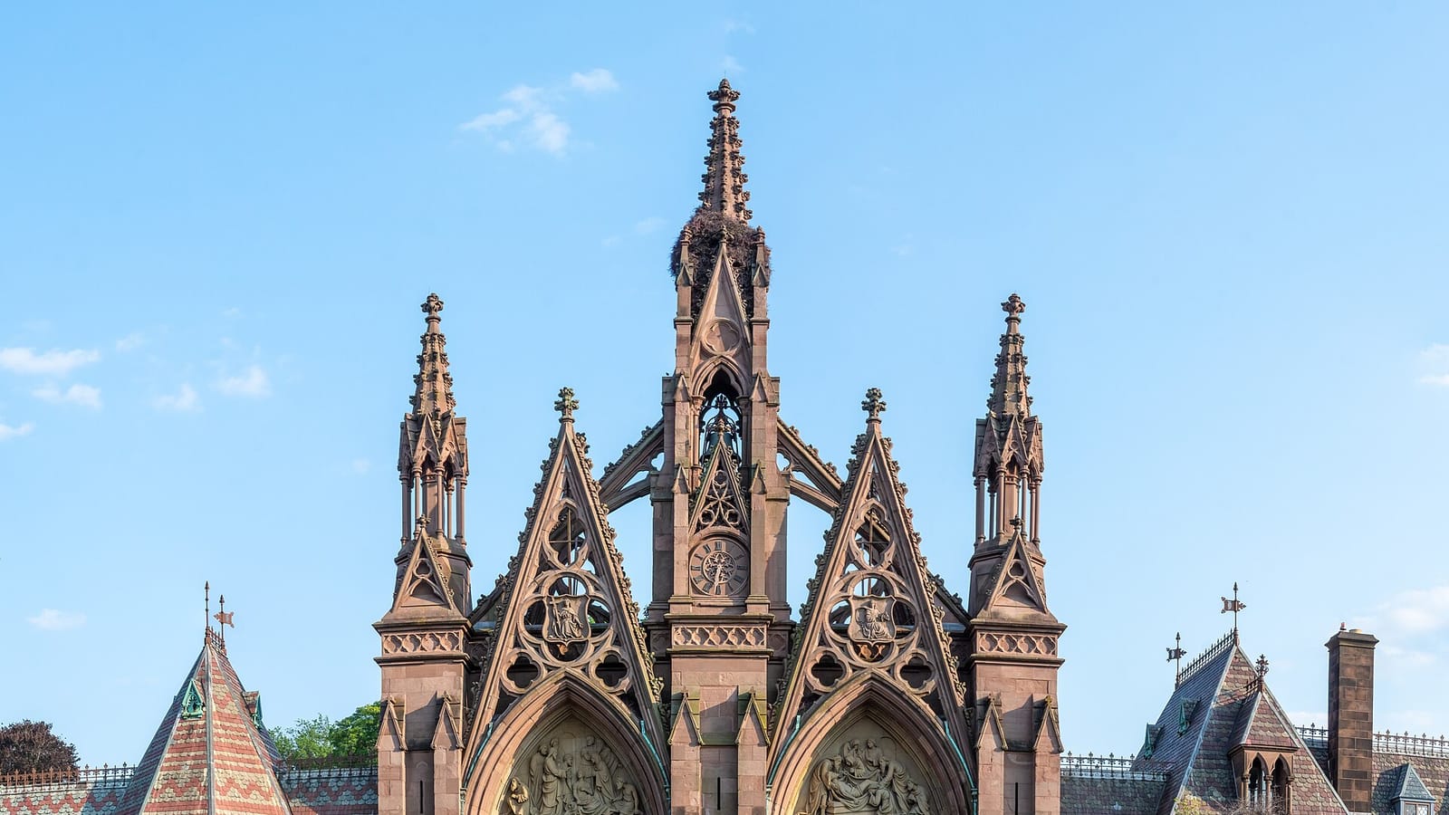 Top of a tall brownstone Gothic gateway with three pointed gables, carved spires, a clock in the central tower and a bird nest near its peak, flanked by patterned slate roofs, under a clear blue sky
