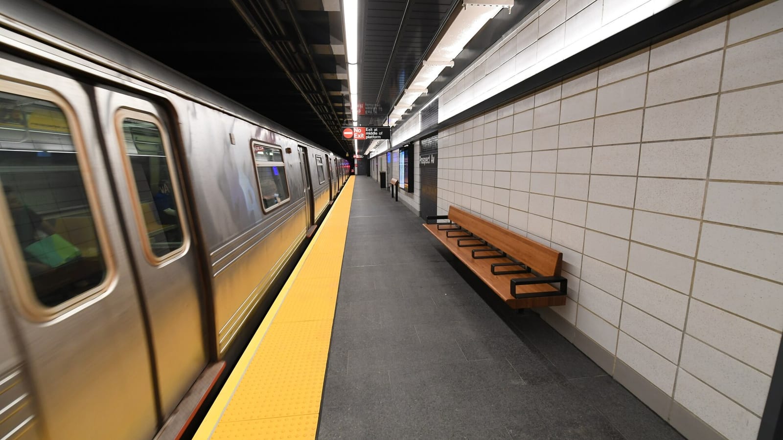 Subway platform with a silver train moving past on the left, a yellow tactile edge strip, a wooden bench against a wall of pale stone tiles, a Prospect Av name panel and a No Exit sign hanging from the ceiling