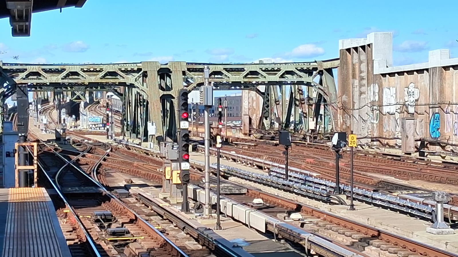Photograph from an elevated subway platform of many tracks and switches, red signal lights, green steel truss structures crossing overhead, and concrete walls with graffiti under a blue sky
