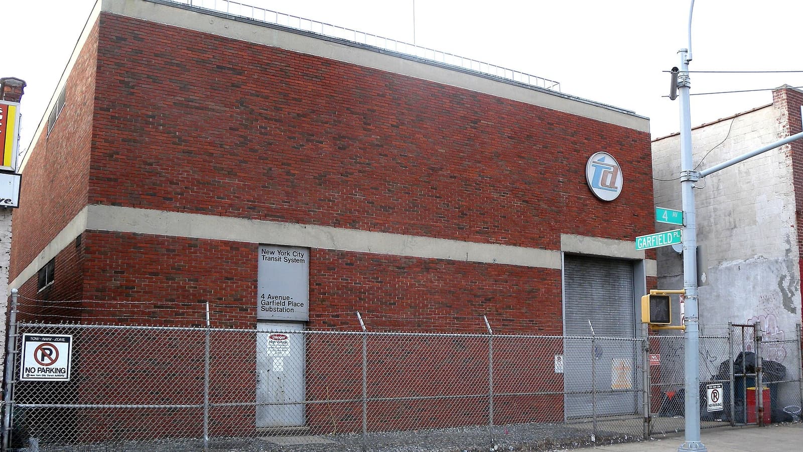 Photograph of a two-story red brick New York City Transit System building marked 4 Avenue-Garfield Place Substation, with a round transit logo, a rolling gray door, a chain-link fence with No Parking signs, and green street signs for 4 Av and Garfield Pl on a pole at right