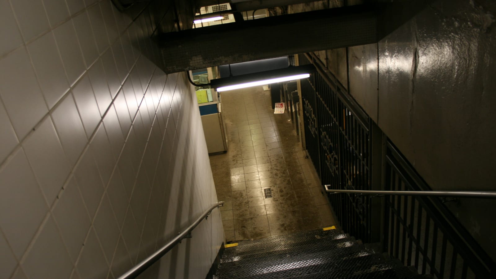 Photograph looking down a steep subway stairway with metal treads, white wall tile, steel handrails, a fluorescent light fixture, and a tiled mezzanine floor below with a token booth and gates
