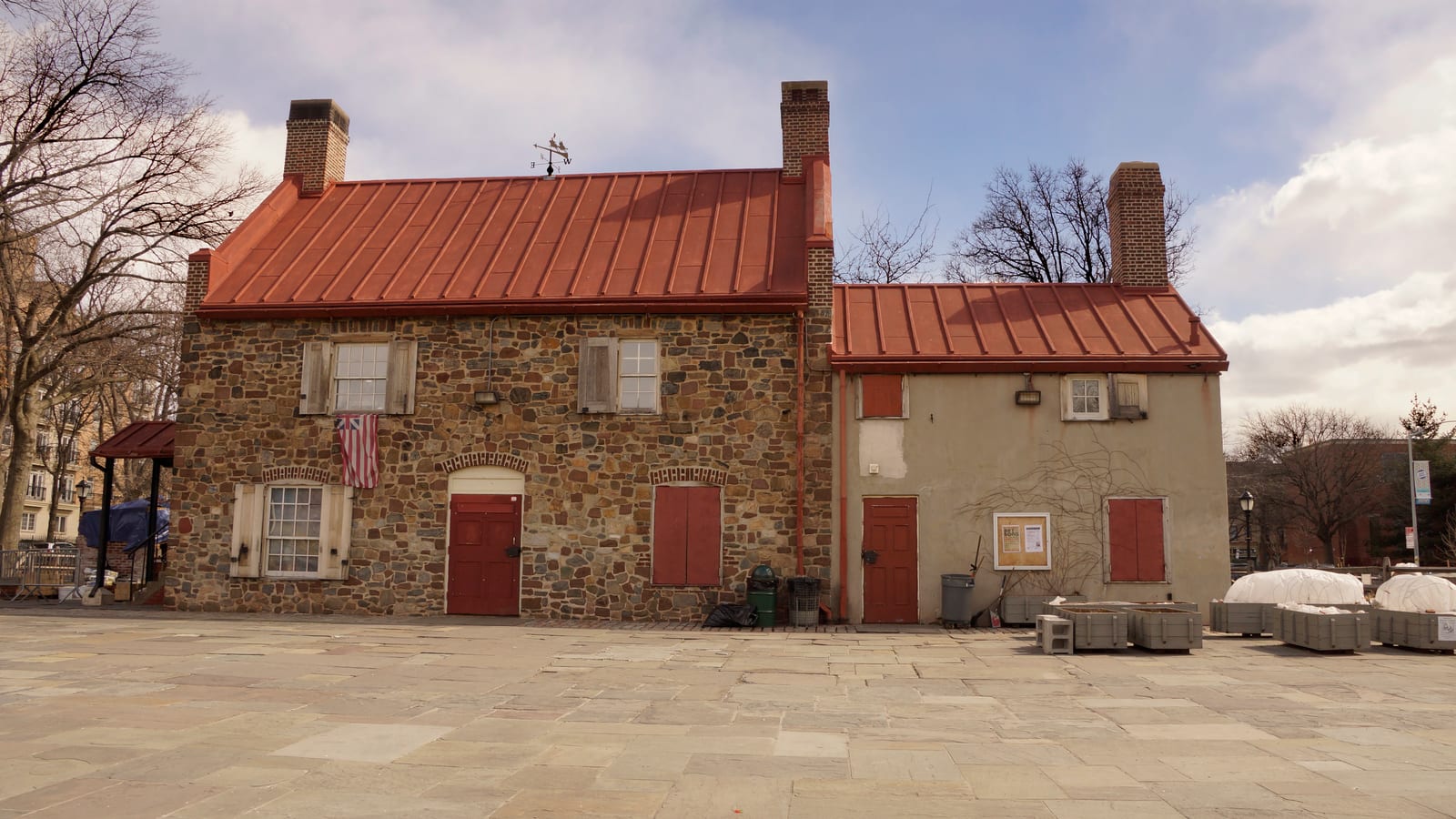 Photograph of a two-story fieldstone house with a red metal roof, brick chimneys, red shutters and doors, and a historic flag by a window, joined to a plaster wing, on a stone-paved plaza with bare trees and covered planters