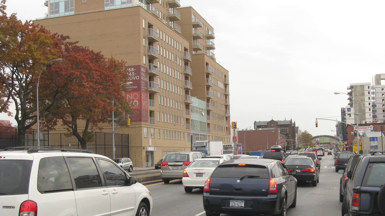 Photograph of a multi-lane avenue with cars in traffic, a tan brick apartment building with balconies and a red NOVO banner at left, autumn trees, traffic lights, and low brick buildings and an elevated train crossing in the distance under a gray sky