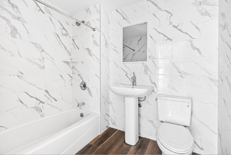 A bathroom with white marble-look wall tile and a dark wood-look floor, a bathtub with a shower rod, a pedestal sink, a mirror and a toilet