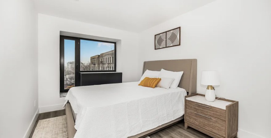A bedroom with gray wood-look floors, a made bed with a beige headboard, a nightstand and lamp, and a dark wall unit under a wide window with older apartment buildings outside