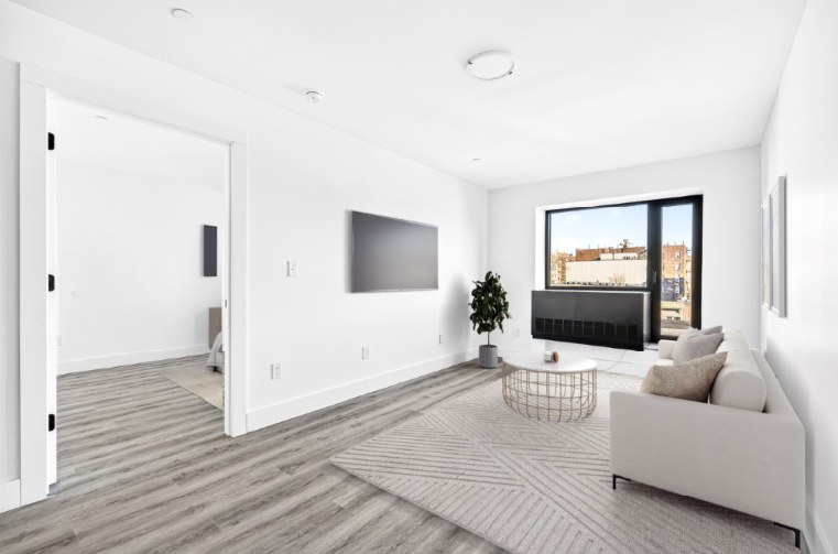 A furnished living room with gray wood-look floors, a white sofa, a round table, a wall-mounted screen that is switched off, a wide window with a dark wall unit under it, and an open door to a bedroom