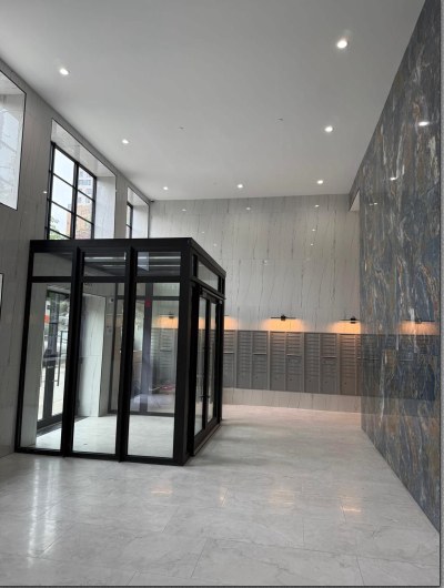 A lobby with tall white stone-look walls and a pale stone floor, a black-framed glass entry vestibule and a wall of mailboxes beyond it