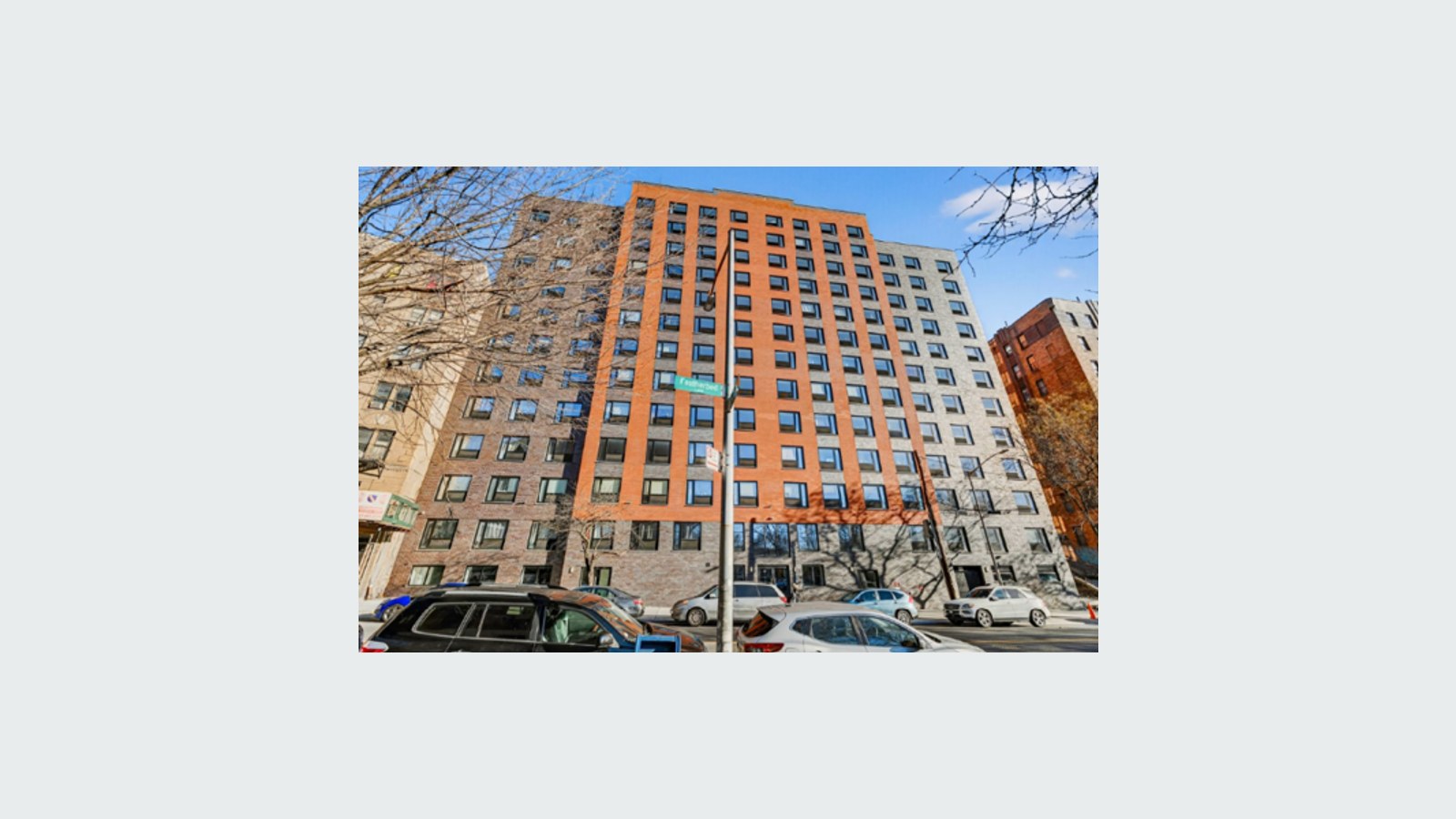 Photograph of a new 12-story apartment building of orange and gray brick with rows of square windows, seen from across the street on a sunny day, with bare tree branches, parked cars, a street light and a green street sign reading Featherbed Ln